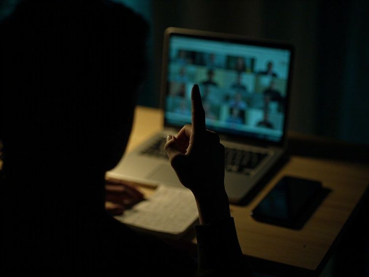 昏暗的夜里，一个人坐在笔记本电脑前，手指竖起指向屏幕上的一格格视频会议画面
