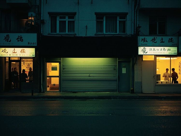 夜晚中国街边的一排小店，中间一家卷帘门半降，隔壁还亮着灯